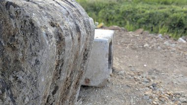 Bir çakıl ve toprak şantiye üzerinde dinlenen kazıcı kovasının aşırı derecede yıpranmış ve desenli tarafında aşırı yakın çekim. Arkasında çimenler olan beton bir blok yanında duruyor..