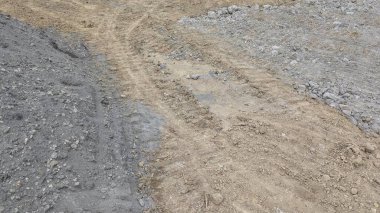 The image shows a construction site with gray gravel on the left right of a central path of light brown dirt. Tire marks are visible on the disturbed earth.