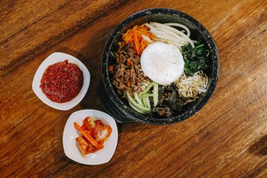 Bibimbap ya da Dolsot olarak bilinen Kore mutfağının düz görüntüsü çiğ yumurta, çiğ et ve acı biber soslu çeşitli sebzelerden oluşan pirinç ve ahşap masadaki kimchi karışımı.