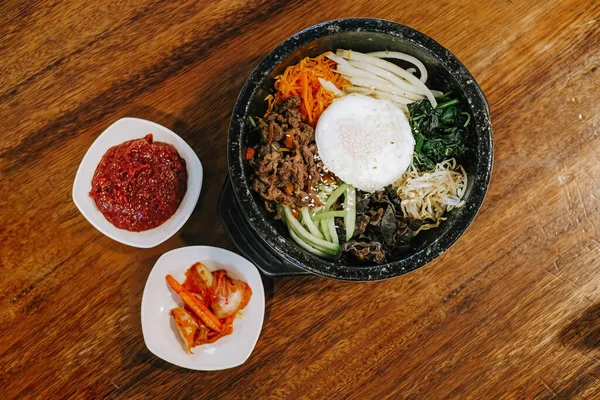 stock image Flat lay or top view shot of Korean cuisine that known as Bibimbap or Dolsot consisting of rice with raw egg, raw meat and assorted vegetables with additional chili sauce and kimchi in a hot plate and wooden table