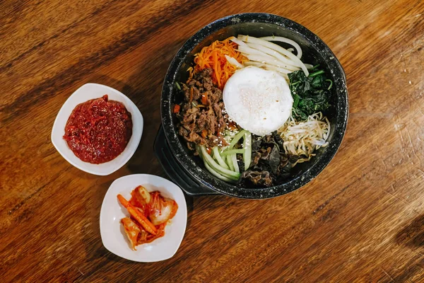 Stock image Flat lay or top view shot of Korean cuisine that known as Bibimbap or Dolsot consisting of rice with raw egg, raw meat and assorted vegetables with additional chili sauce and kimchi in a hot plate and wooden table