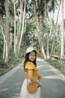 Güneydoğu Asyalı bir kızın yüksek çözünürlüklü fotoğrafı baharatlı hardal gömleği, beyaz etek, turta çantası ve yolun ortasında palmiye ağaçlarıyla çevrili bir şapka gibi duran tropik bir şapka giyer.