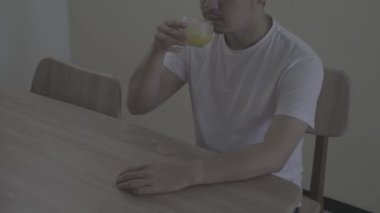 Beyaz gömlek giyen genç bir adam ahşap sandalyeye oturur ve tahta masada bir bardak portakal suyu içer.