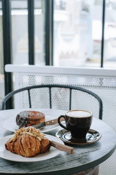 Portrait or Vertical Shot of Breakfast Set: Almond Croissant, Chocolate Crombolomi, and a Cup of Coffee on Wooden Table. Focus on the Cup of Coffee.