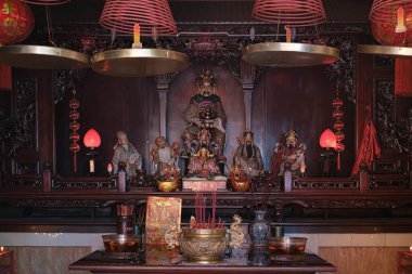 İçinde birkaç Çin tanrısı heykeli ve kırmızı ve sarı süslemelerle çevrili mumlar olan kapalı bir mabet. Ayrıca bir raf dolusu altın kase, vazo ve lamba var. Bu resimdeki baskın renkler siyah, kahverengi ve kırmızıdır..