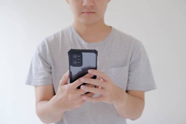 Gri t-shirt giyen Asyalı genç bir adamın orta boy resmi elinde ve akıllı telefona bakıyor. Telefona odaklanmış. Sığ alan derinliği. İzole edilmiş beyaz arkaplan.