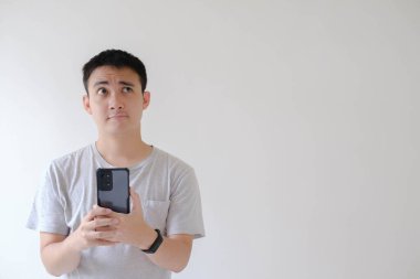 Gri tişörtlü ve akıllı gözlüklü Asyalı genç bir adam akıllı telefonu tutuyor ve kafası karışmış bir şekilde ona bakıyor. İzole edilmiş beyaz arkaplan.