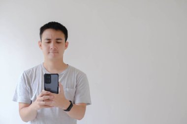 Gri tişörtlü ve akıllı gözlüklü Asyalı genç bir adam elinde tutuyor ve kafası karışmış bir şekilde akıllı telefona bakıyor.