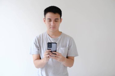 Gri tişört giyen genç bir Asyalı 'nın orta açı ve yumuşak odaklı fotoğrafı akıllı bir telefona bakıyor. Sığ alan derinliği. İzole edilmiş beyaz arkaplan.