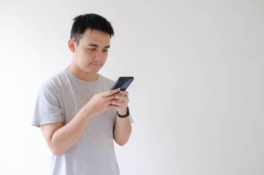 Gri tişört giyen ve bileğinde akıllı kaset olan Asyalı genç bir adam akıllı telefonu tutuyor ve ona bakıyor. İzole edilmiş beyaz arkaplan.