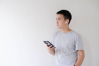 Gri tişört giyen genç bir Asyalı adam, bir çift kablosuz kulaklık takıyor, ve bir akıllı telefon tutuyor ve sol tarafına bakıyor. İzole edilmiş beyaz arkaplan.
