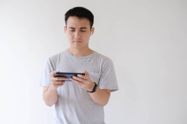 Gri tişört giyen genç bir Asyalı akıllı telefonda kafası karışık bir ifadeyle oyun oynuyor. İzole edilmiş beyaz arkaplan.