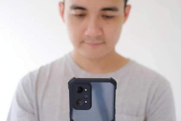一位身穿灰色T恤的年轻亚洲男子的部分脸的特写镜头正在拿着并在看一部智能手机 集中在电话上 浅浅的野战深度孤立的白色背景 — 图库照片