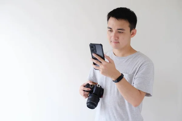 Gri tişört giyen genç bir Asyalı sol elinde akıllı bir telefona bakıyor ve sağ elinde bir DSLR kamerası tutuyor. İzole edilmiş beyaz arkaplan.