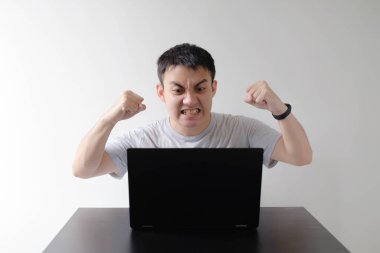 Gri tişört giyen genç bir Asyalı ve sol bileğindeki akıllı bir adam siyah bir masanın üzerinde duran siyah bir dizüstü bilgisayarın önünde sıkışan yumruklara kızar. İzole edilmiş beyaz arkaplan.