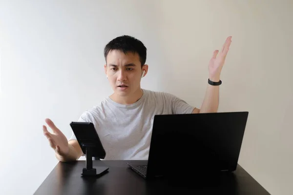 A young Asian man wearing a gray t-shirt, a smartwatch on his left wrist, and a pair of wireless earphones is angry at an online meeting. Laptop and phone on wooden table. Isolated white background.