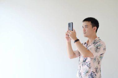Tropikal desenli pembe gömlek giyen genç bir Asyalı ve sol bileğinde bir akıllı telefon kullanarak fotoğraf çekiyor ya da çekiyor. İzole edilmiş beyaz arkaplan.