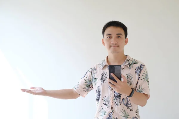 一位年轻的亚洲男子穿着一件粉色衬衫 图案是热带的 左手腕上戴着一顶漂亮的图章 他的左手拿着智能手机 右手是张开的手掌 孤立的白色背景 — 图库照片