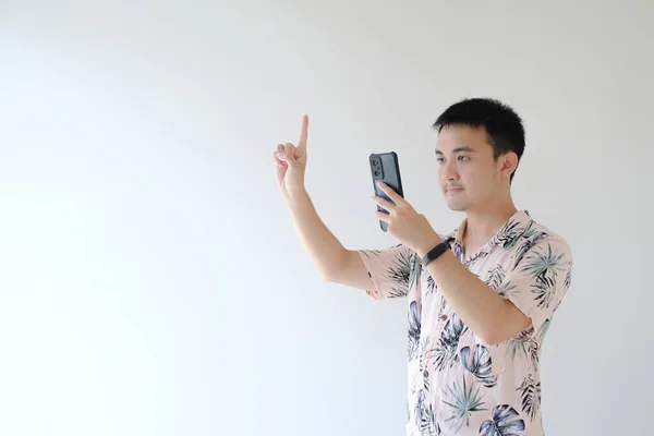 stock image A young Asian man wearing a pink shirt with tropical patterns and a smartwatch on his left wrist is photographing using a smartphone and his finger indicated one. Isolated white background.