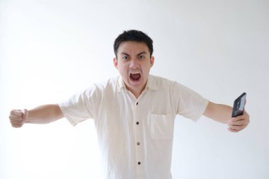 Bej tişört giyen genç bir Asyalı ve sol bileğinde akıllı bir bant var. Kameraya öfkeyle bakıyor ve elinde akıllı bir telefon tutuyor. İzole edilmiş beyaz arka plan. Reklam için uygun..