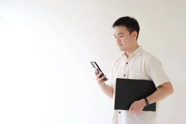 stock image A young Asian man wearing a beige shirt and a smartwatch on his left wrist is carrying a black laptop while holding and looking at his smartphone. Isolated white background. Suitable for advertisement.