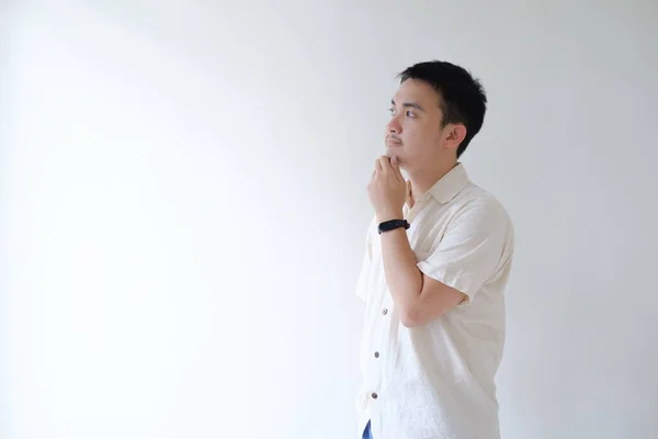 stock image A young Asian man wearing a beige shirt and a smartwatch is thinking of putting his thumb and pointer fingers to his chin. Isolated white background. Negative space.