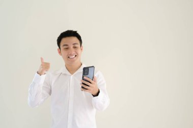 Beyaz tişörtlü genç Asyalı bir adam gülümseyen bir yüz ifadesiyle akıllı telefonu tutarken başparmağını kaldırıyor. İzole edilmiş beyaz arkaplan.