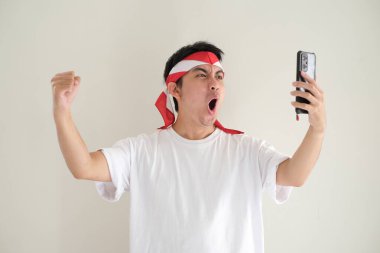 Beyaz tişört ve Kemerdekaan Endonezya kostümü giyen Asyalı genç bir adam akıllı telefonuna bakarken heyecanlı bir ifade hissediyor. Endonezya Bağımsızlık Günü. İzole edilmiş beyaz arkaplan.