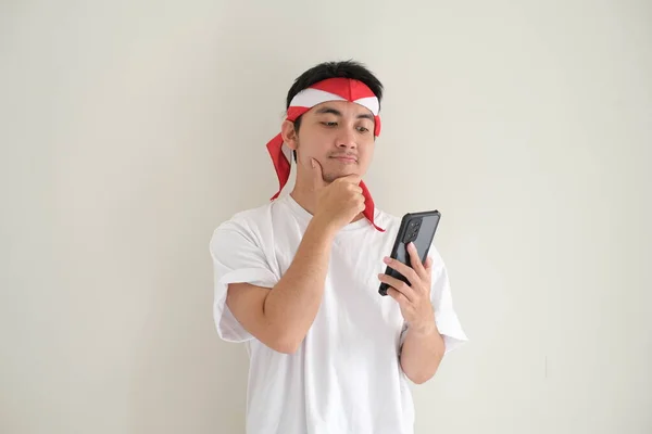 Beyaz tişört ve Kemerdekaan Endonezya kostümü giyen genç bir Asyalı akıllı telefonuna bakarken düşünüyor. Endonezya Bağımsızlık Günü. İzole edilmiş beyaz arkaplan.