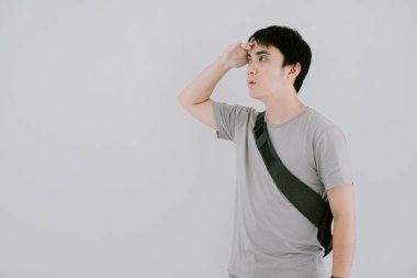 Asyalı genç bir adamın adaçayı yeşili günlük tişört giydiği seçici odak fotoğrafı, akıllı göz bandı ve minimalist askı çantası, izole edilmiş beyaz arka planda, el üstünde, çok uzaklardan bakıyor.