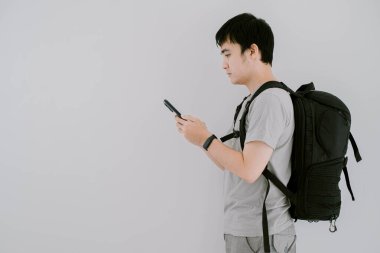 Asyalı genç bir adamın seçici odak noktası. Adaçayı yeşili, sıradan tişört, akıllı sırt çantası ve akıllı sırt çantası giyerken sol tarafta izole edilmiş beyaz arka planda akıllı telefona bakıyor.