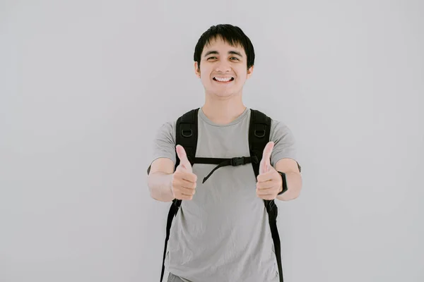 Asyalı genç bir adamın adaçayı yeşili bir tişört giydiği seçici odak fotoğrafı, akıllı kollu tişört ve gülücük ifadeli sırt çantası, izole edilmiş beyaz arka planda kameraya çift parmak veriyor.