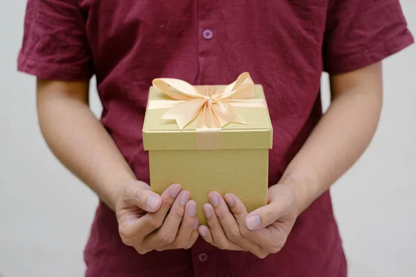 Male hands holding a small gold gift box wrapped with gold ribbon. Close-up and indoor shot. Selective focus. Christmas Concept.