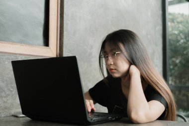Günlük kıyafet giyen genç Asyalı kadın endüstriyel bir odada ya da kafede stresli bir dizüstü bilgisayar üzerinde çalışıyor.