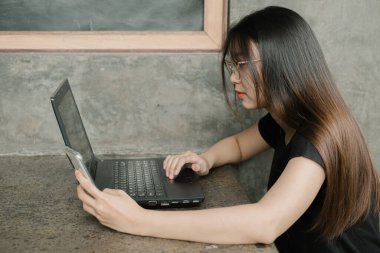 Günlük kıyafet giyen genç Asyalı kadın endüstriyel bir odada ya da kafede dizüstü bilgisayarla çalışıyor.