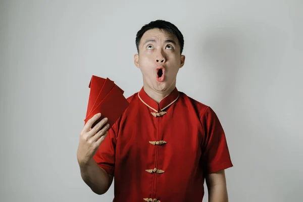 Shocked face of Asian man wearing Cheongsam or Chinese traditional cloth while fanning his self using angpao or red monetary gift on white background. Gong Xi Fa Cai.