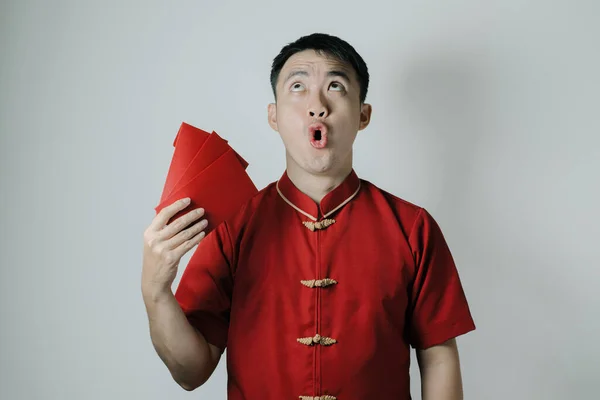 Shocked face of Asian man wearing Cheongsam or Chinese traditional cloth while fanning his self using angpao or red monetary gift on white background. Gong Xi Fa Cai.