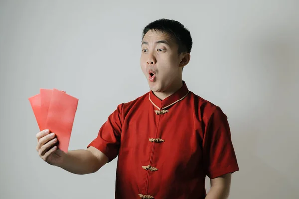 Shocked face of Asian man wearing Cheongsam or Chinese traditional cloth while holding angpao or red monetary gift on white background. Gong Xi Fa Cai.
