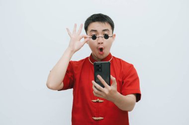 Beyaz arka plandaki akıllı telefona bakarken Cheongsam ve siyah gözlüklü Asyalı adamın şok olmuş yüzü.