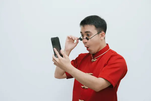 Shocked face of Asian man wearing Cheongsam and black vintage sunglasses while looking at the smartphone on white background