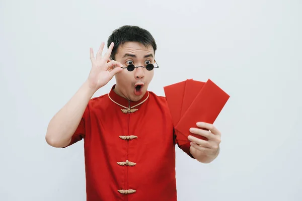 Wow face of Asian man wearing Cheongsam and black rounded vintage sunglasses while holding and looking at the angpao or red monetary gift on white background. Chinese New Year concept. Gong Xi Fa Cai.
