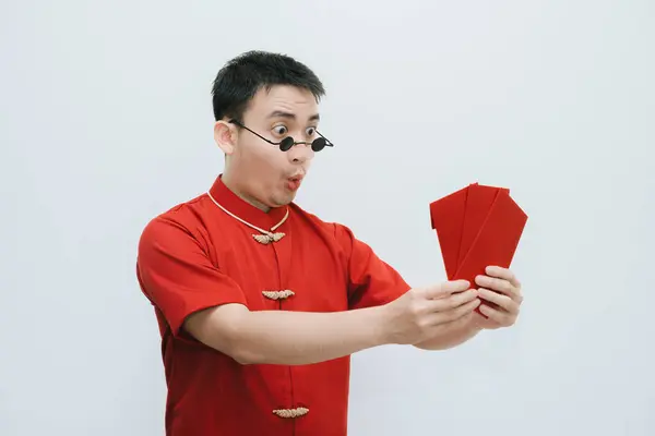 Shocked face of Asian man wearing Cheongsam and black rounded vintage sunglasses while holding and looking at the angpao or red monetary gift on white background. Gong Xi Fa Cai.