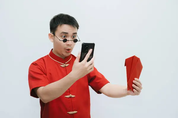 Shocked face of Asian man wearing Cheongsam and black rounded vintage sunglasses while holding at the angpao or red monetary gift and looking at his smartphone on white background. Gong Xi Fa Cai.