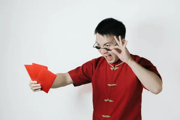 Surprised face of Asian man wearing traditional costume holding angpao or red monetary gift on white background. Chinese New Year concept. Gong Xi Fa Cai.