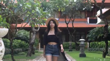 Asyalı bir kadın rattan şapka takıyor ve Bali tatil köyünde yürüyor. Seyahat görüntüleri. Ağır çekim. Her yere git.