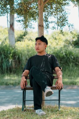 Genç Güneydoğu Asyalı bir adamın portresi ya da dikey portresi ağacın altındaki tahta sandalyede otururken, sol tarafına bokeh arka planıyla bakıyor. Yakın çekim..