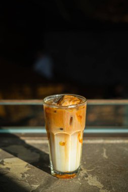 Bir kahve dükkanında, bir bardak buzlu kahve ve sütlü krema. Pencere ve gölgenin kontrastlı güneş ışığı kombinasyonu. Dikey veya dikey çekim.