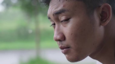 Genç Güneydoğu Asyalı bir adam yağmur sırasında üzgün ve bokeh arkaplanlı kameraya bakıyor. Yavaş çekim..