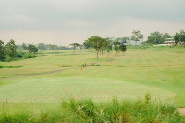 Golf sahasında iyi bakılmış yeşil çimenler ve bulutlu hava koşullarında deliğin işaretlendiği kırmızı bir bayrak var. Tarlalar ağaçlarla çevrili..