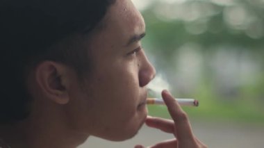 Arka planı bulanık veya bokeh etkisi olan Güneydoğu Asyalı bir adam sigara içiyor. Yavaş çekim görüntüsü.